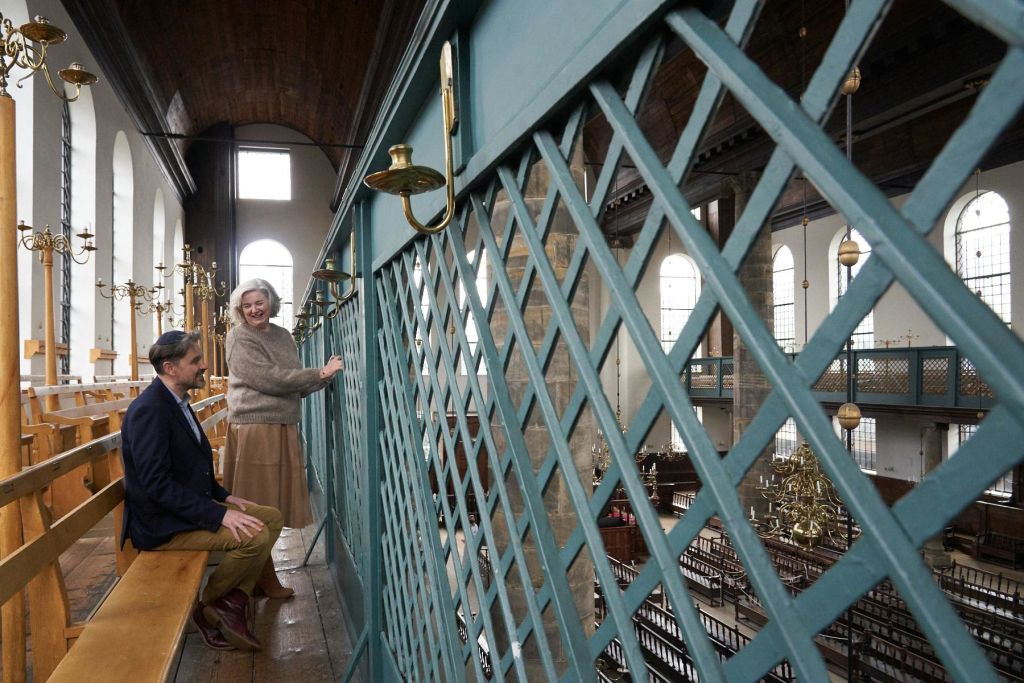 두 사람이 교회 내부의 나무 발코니에서 대화를 나누고 있습니다. 커다란 창문과 화려한 황동 캔들 홀더가 벽을 둘러싸고 있습니다.