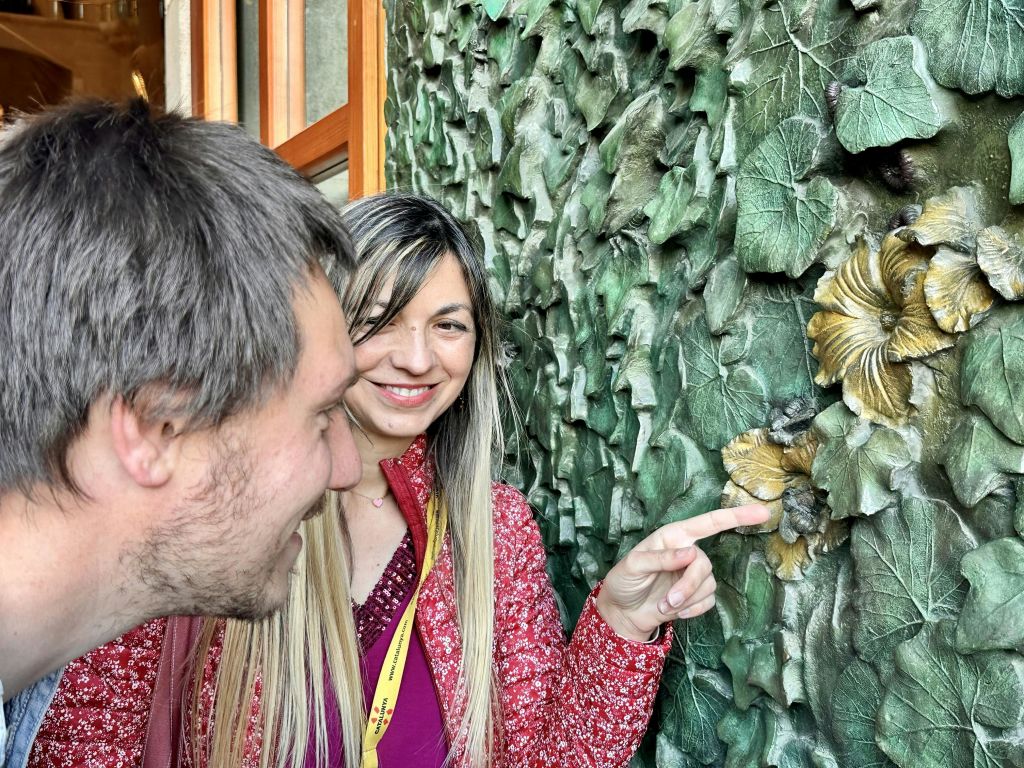 두 사람이 미소를 지으며 질감이 살아있는 초록색 벽을 바라보고 있는데, 여인이 금색의 작은 조각을 가리키고 있습니다.