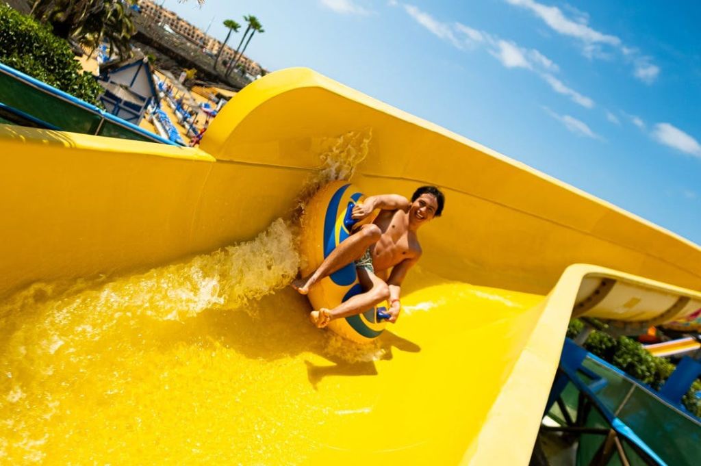 화창한 날 워터파크에서 노란색 워터 슬라이드 아래로 팽창식 튜브를 타고 내려가는 사람.