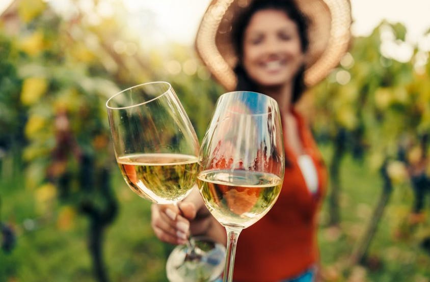 포도원을 배경으로 모자를 쓴 여성이 흐릿하게 웃고 있는 가운데, 화이트 와인 두 잔이 초점을 맞추고 있습니다.
