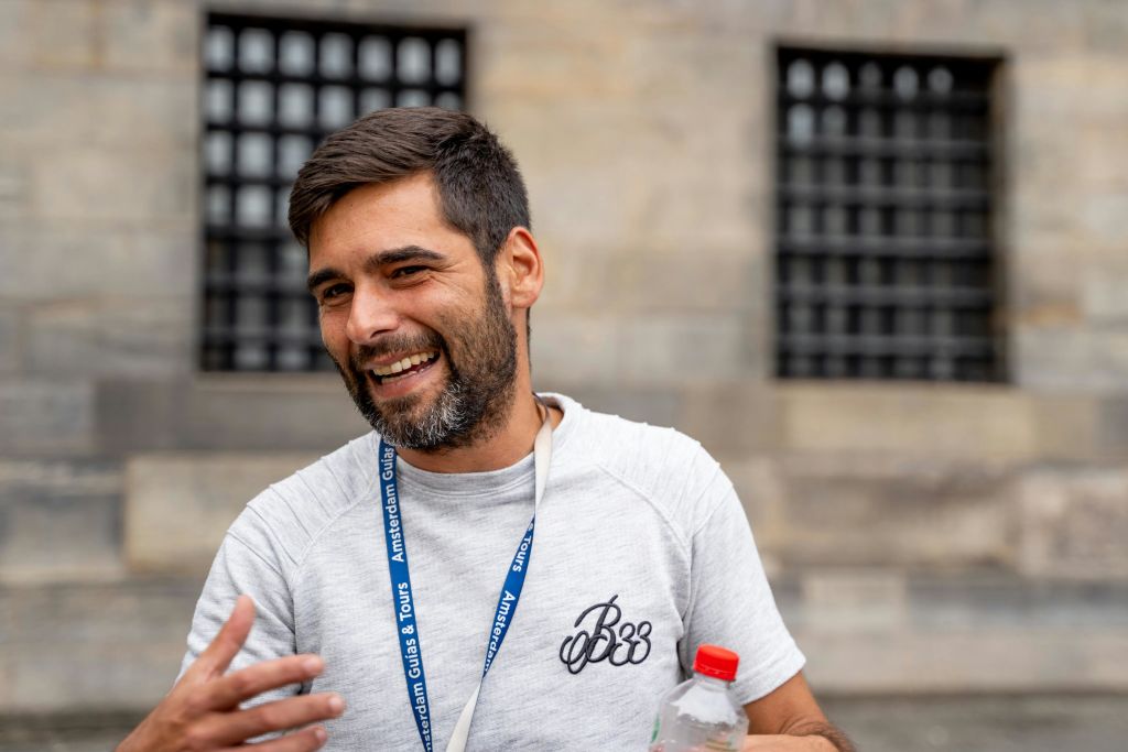 수염과 짧은 머리를 한 남자가 물병을 들고 미소를 짓고 있습니다. 그는 랜야드를 착용하고 석조 건물 앞에 서 있습니다.