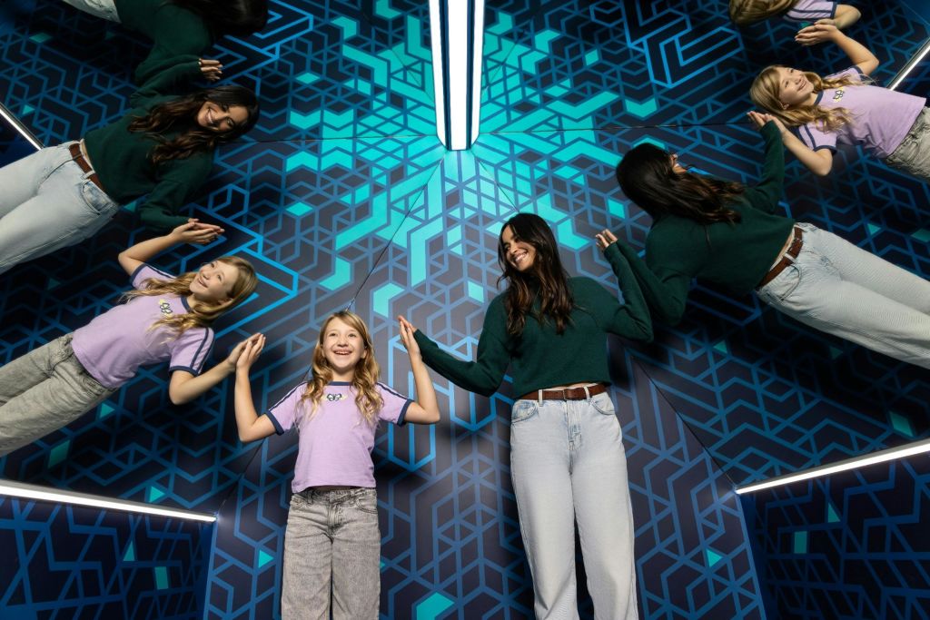 보라색 셔츠를 입은 어린이와 초록색 스웨터를 입은 성인이 기하학적 거울 벽 앞에 서 있습니다.