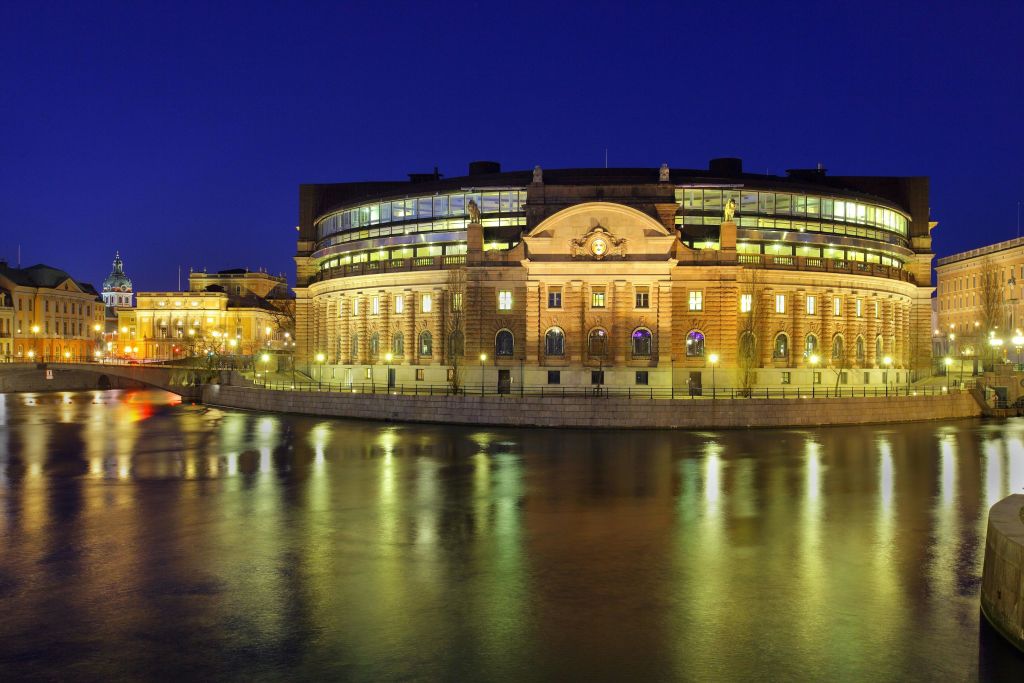 아치형 창문과 현대적인 유리 섹션이 있는 조명으로 장식된 역사적인 건물로, 밤에는 잔잔한 수로에 비춰집니다.