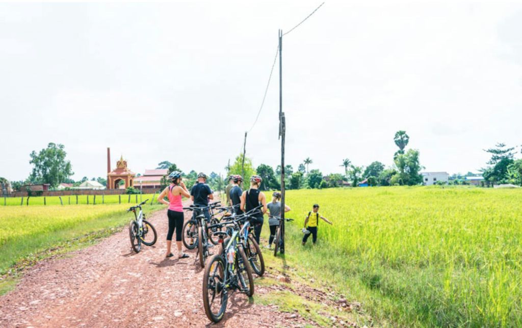 녹색 밭 옆의 흙길에서 자전거를 타고 건물이 보이는 배경을 배경으로 한 무리의 자전거 운전자들이 서 있습니다.