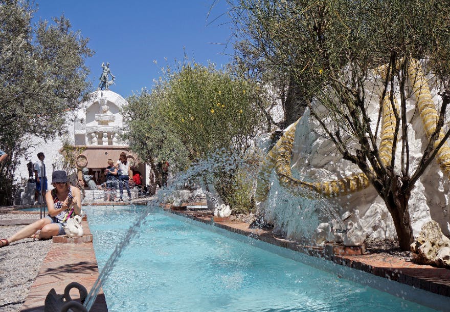 푸른 하늘 아래 녹음과 장식용 뱀 조각품으로 둘러싸인 청록색 분수 근처에 사람들이 모여듭니다.