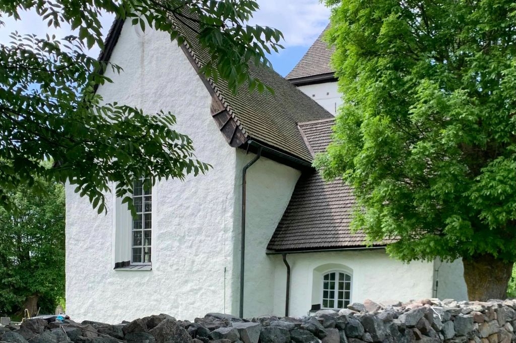 전경의 푸른 나무와 바위에 의해 부분적으로 가려진 가파른 갈색 지붕이 있는 흰색 건물입니다.