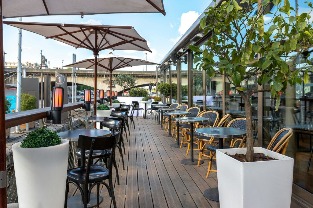 나무 데킹, 테이블, 의자, 우산, 화분, 다리를 배경으로 한 전망을 즐길 수 있는 야외 카페.