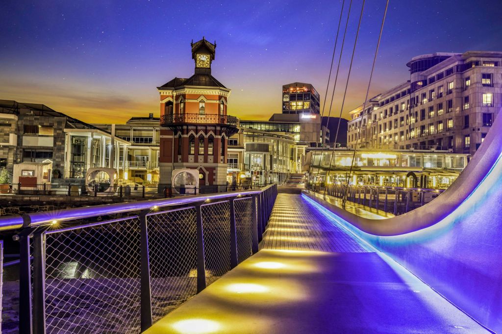 별이 빛나는 하늘을 배경으로 시계탑과 현대적인 건물이 있는 밤에 조명이 켜진 보행자용 다리입니다.