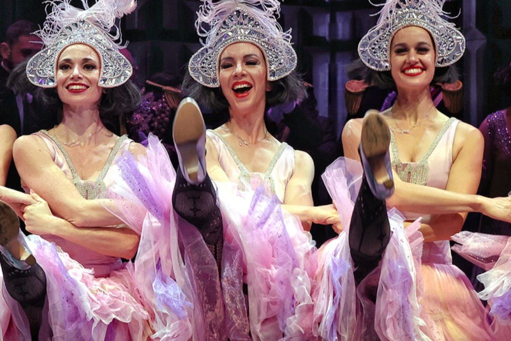 분홍색 의상을 입은 두 명의 공연자와 정교한 헤드피스가 무대에서 춤을 추며 일제히 다리를 걷어차고 있습니다.