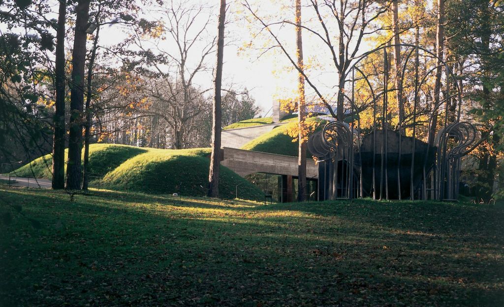 초록색 잔디로 덮인 지붕이 있는 현대적인 건물은 가을에 키가 크고 맨발로 나무가 우거진 숲 풍경과 어우러집니다.