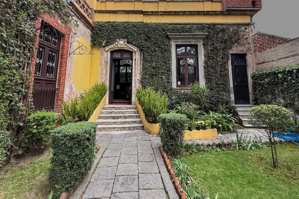 담쟁이가 덮인 벽과 창문, 입구로 이어지는 작은 계단이 있는 노란색 건물로 이어지는 돌길이 있는 마당입니다.