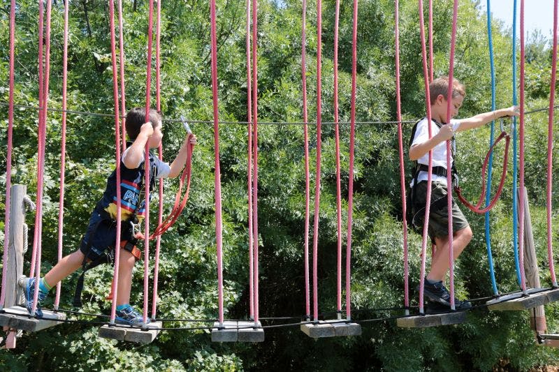 빽빽한 녹색 단풍으로 둘러싸인 붉은 밧줄과 플랫폼이 있는 밧줄 다리를 걷고 있는 두 어린이. 둘 다 하네스를 착용합니다.