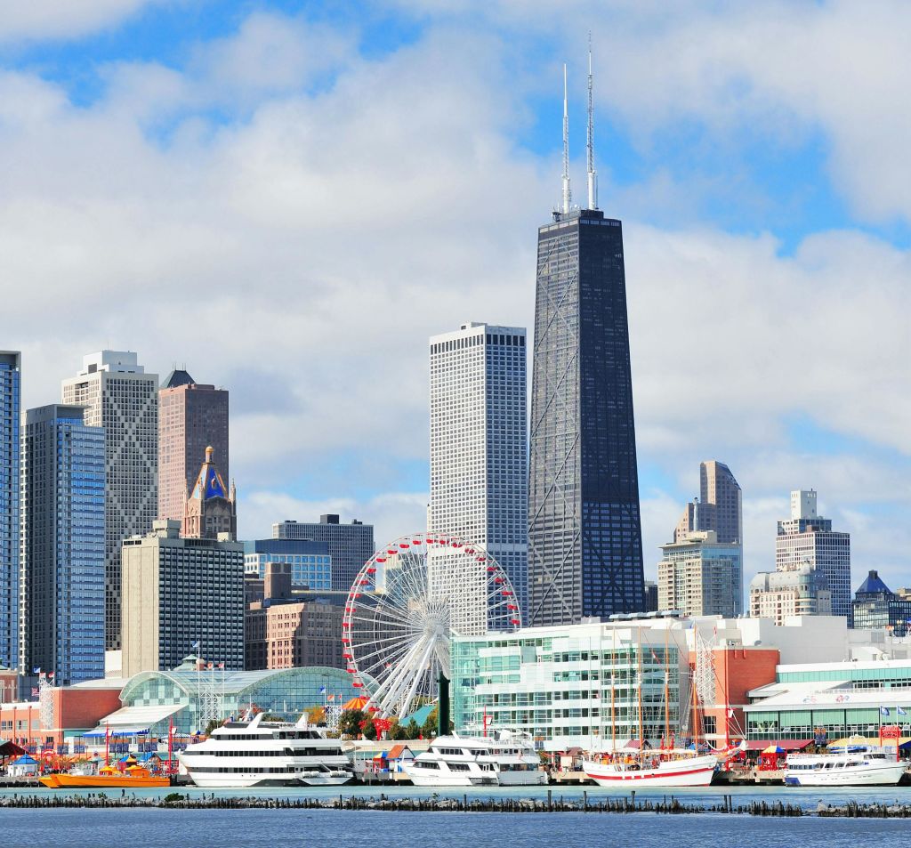 높은 건물과 대관람차, 그리고 부분적으로 흐린 하늘 아래 바닷가에 정박한 여러 보트가 있는 도시 스카이라인입니다.