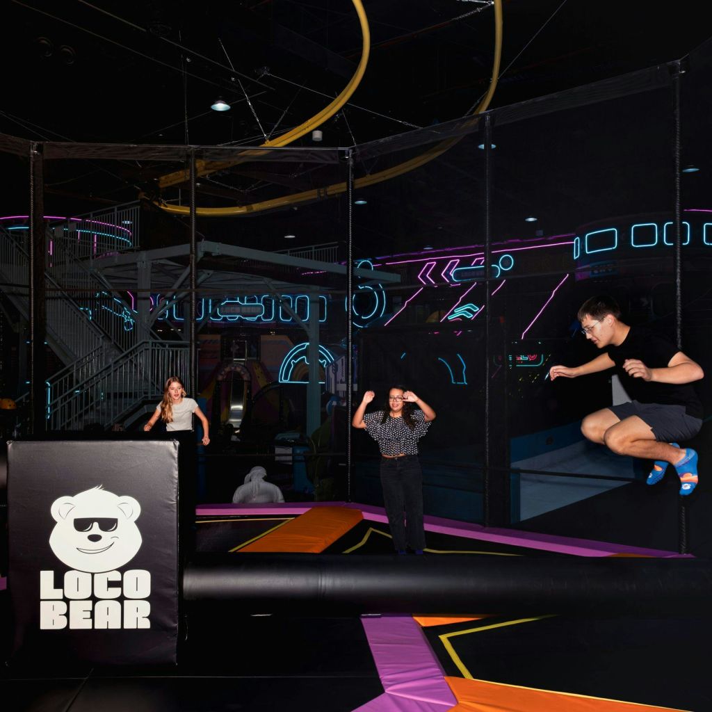 네온 조명 실내 트램펄린 공원 안에 세 사람이 있습니다. 한 사람은 점프하고 다른 두 사람은 일어섭니다. "Loco Bear" 로고가 표시됩니다.