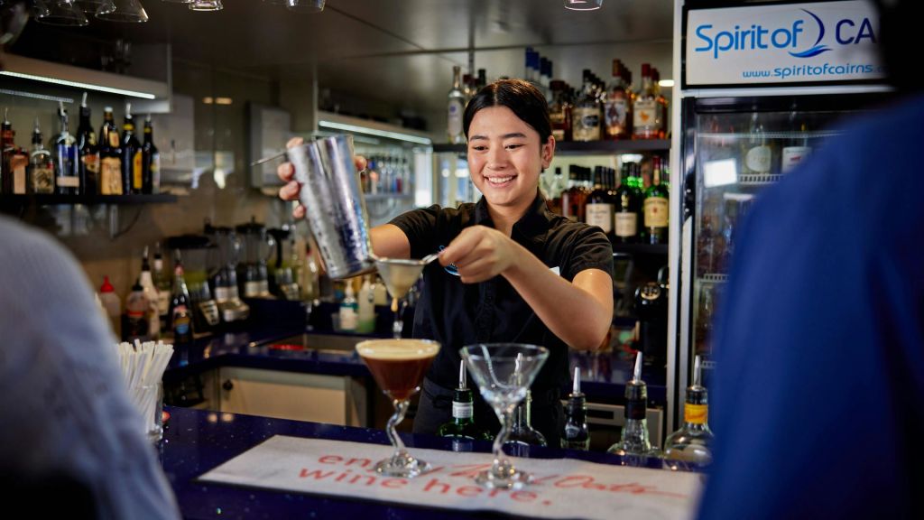 다양한 병을 배경으로 한 바에서 셰이커의 칵테일을 잔에 부으며 웃고 있는 바텐더.