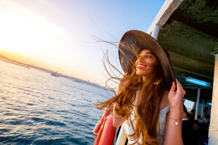 바람이 머리카락을 날리면서 물과 스카이라인을 배경으로 활짝 핀 모자를 들고 보트에서 미소를 짓고 있는 여성.