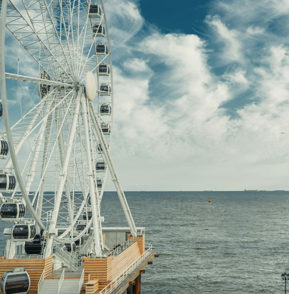나무 부두 옆에 흰색 곤돌라가 있는 대형 관람차로, 잔잔한 바다와 흩어진 구름이 있는 하늘이 내려다보입니다.
