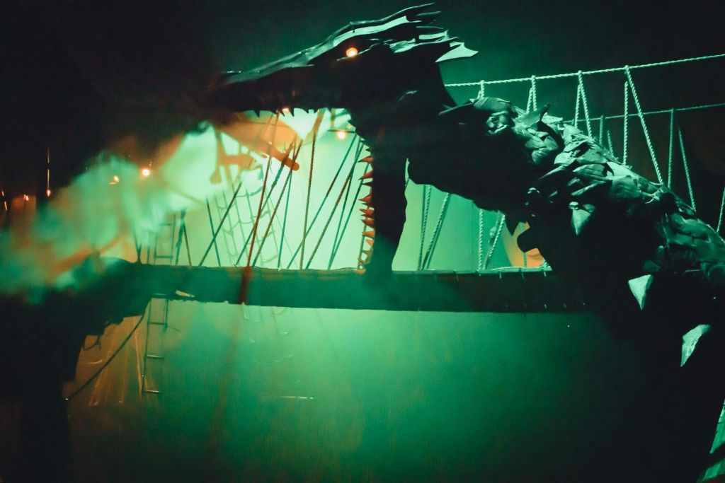 안개가 자욱한 현수교에서 빛나는 눈을 가진 커다란 기계식 드래곤이 녹색과 주황색 조명으로 빛납니다.