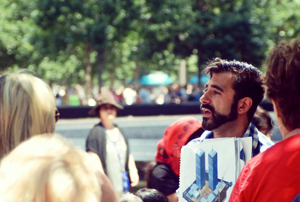 수염을 기른 남성이 프린트된 쌍둥이 빌딩을 배경으로 흐릿한 나무가 우거진 야외에 서 있습니다.