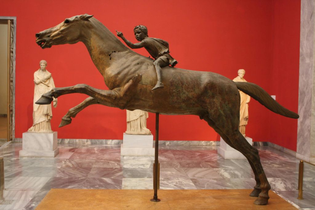 붉은 벽과 대리석 바닥이 있고 다른 조각상으로 둘러싸인 박물관에 전시된, 말을 타고 있는 아이의 청동 동상입니다.