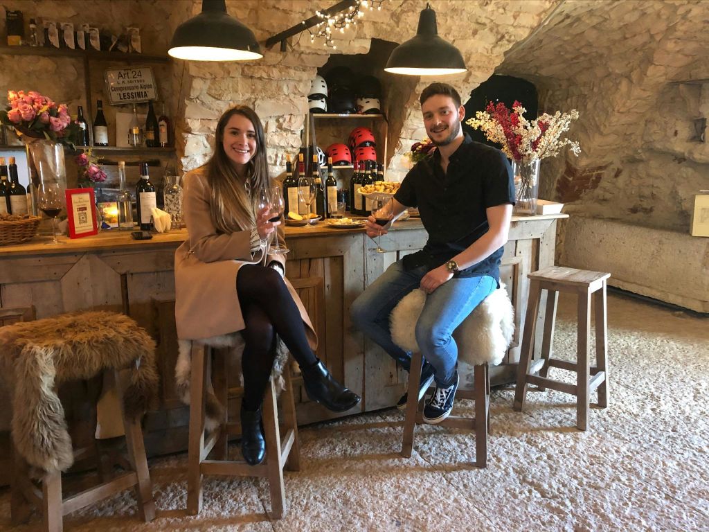 돌담, 병, 따뜻한 조명이 있는 소박하고 아늑한 바에서 바스툴에 앉아 와인을 즐기는 두 사람.