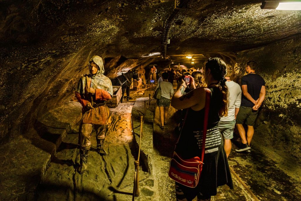 역사적인 옷을 입은 광부의 동상이 있는 어두운 지하 터널을 걷는 사람들. 일부는 사진을 찍고 있습니다.