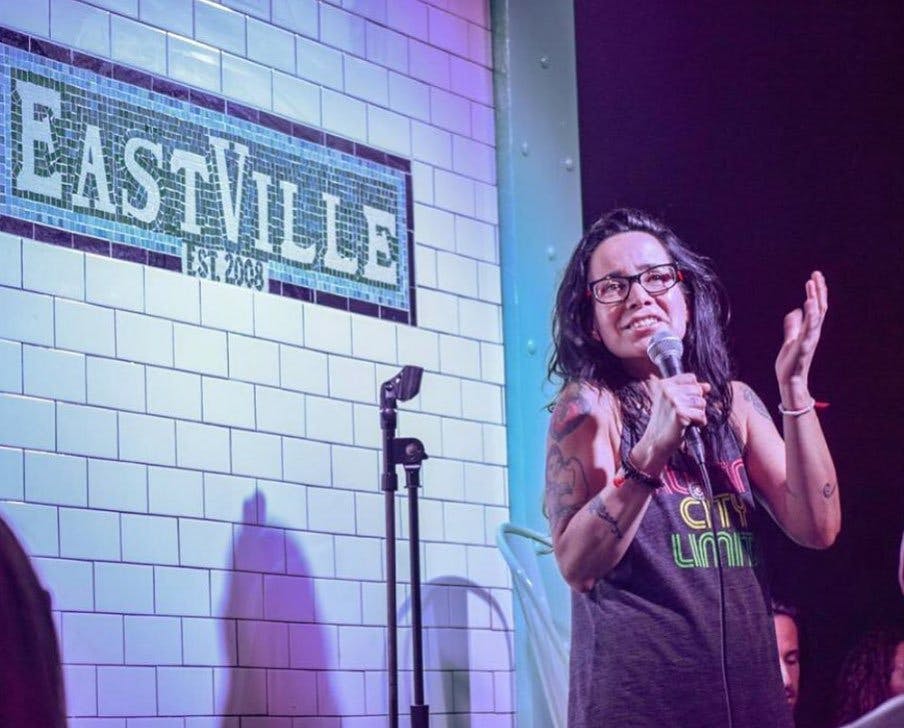 무대에서 마이크를 들고 스탠드업 코미디를 하는 안경과 문신을 한 사람. 배경은 "Setville" 표지판이 있는 타일 벽을 보여줍니다.