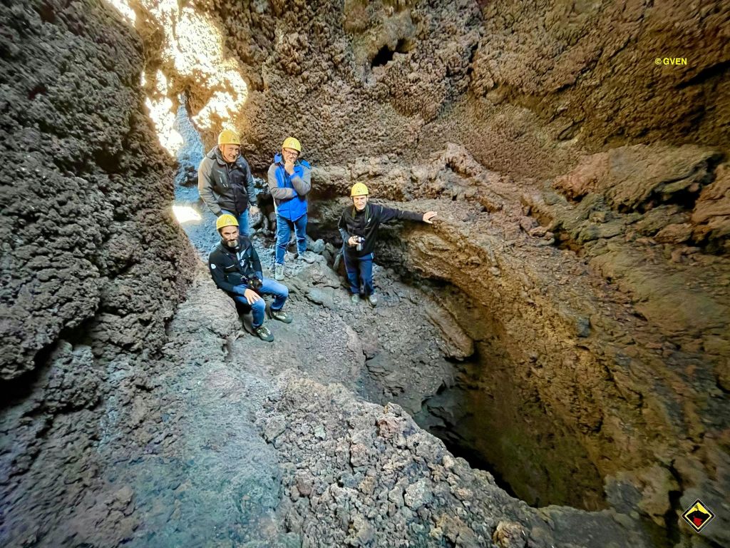 험준한 벽이 있는 바위 동굴 안에는 헬멧을 쓰고 야외 옷을 입은 네 사람이 서 있습니다. 한 사람은 바깥을 향하고 있습니다.