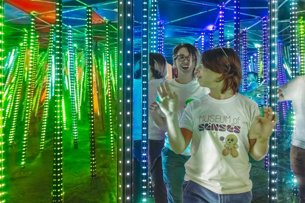 "감각의 박물관" 티셔츠를 입고 미소를 짓고 탐험하는 다채로운 LED 조명이 있는 거울 미로의 두 사람.