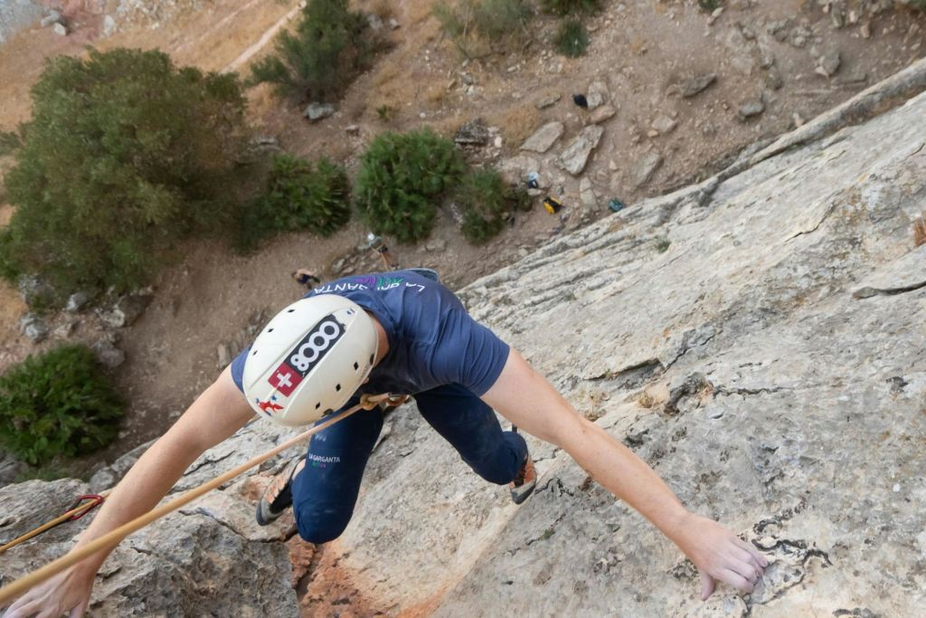 등산객이 헬멧과 마구를 착용한 채 가파른 바위 얼굴로 올라가고, 아래에는 나무와 바위가 배경으로 보입니다.