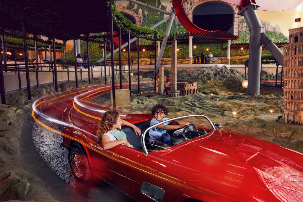 트랙과 롤러코스터를 배경으로 한 미니어처 풍경에 둘러싸인 두 어린이가 빨간색 자동차를 타고 미소를 짓고 앉아 있습니다.