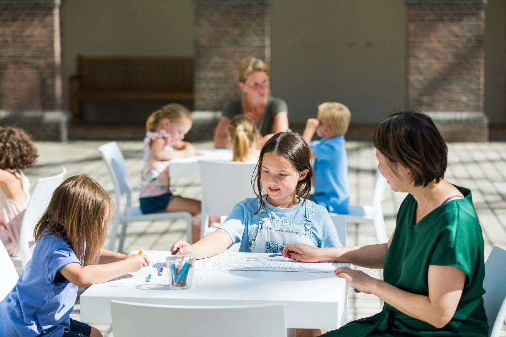 흰색 테이블에 앉아 햇살이 내리쬐는 실내에서 색연필로 그림 그리기 활동을 하는 어린이와 성인.