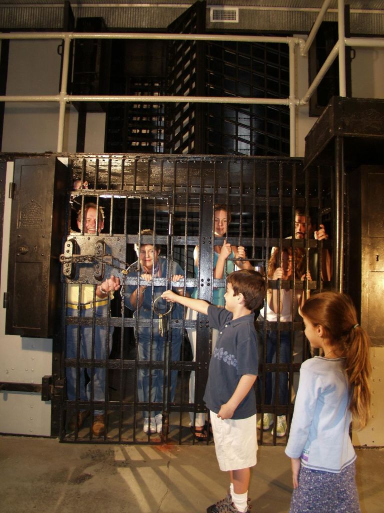 커다란 검은색 감옥 안에 성인 4명과 어린이 1명이 서 있고, 다른 어린이 2명이 열쇠를 들고 밖에 서 있습니다.