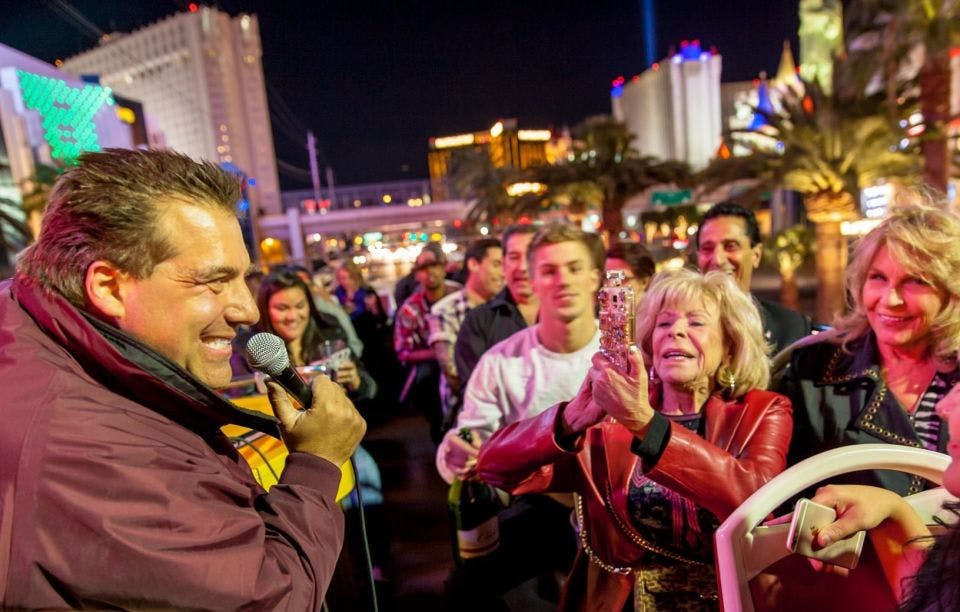 빨간 재킷을 입은 나이가 많은 여성이 사진을 찍는 등 많은 사람들이 밤에 활기차고 밝은 거리에 모여듭니다.