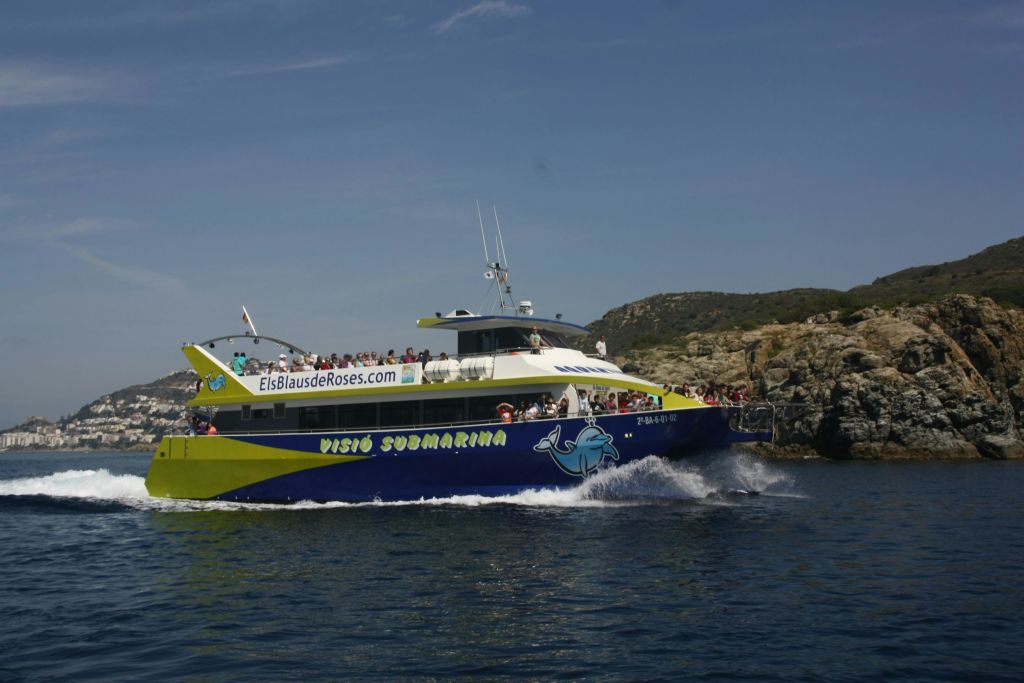 맑은 하늘 아래 바위 해안 근처에서 "Visió Submarina" 와 "ElsBlausdeRoses.com" 이라는 라벨이 붙은 투어 보트.