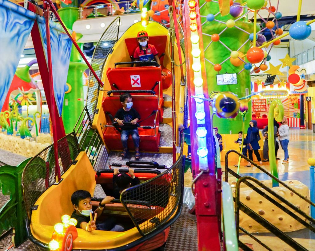 아이들이 마스크를 착용하고 다채로운 색상의 실내 놀이공원 곤돌라를 타고 있습니다. 밝은 조명과 장난기 넘치는 장식이 눈길을 사로잡습니다.