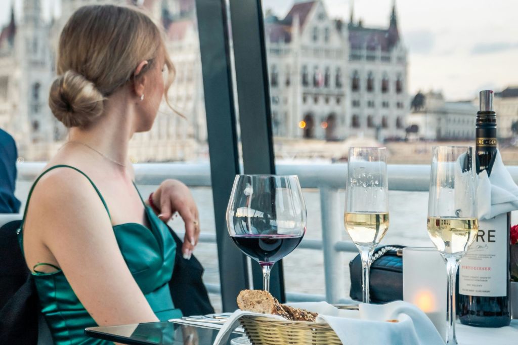 녹색 드레스를 입은 여성이 와인잔과 빵이 놓인 테이블에 앉아 물 건너편 건물을 내려다보고 있습니다.