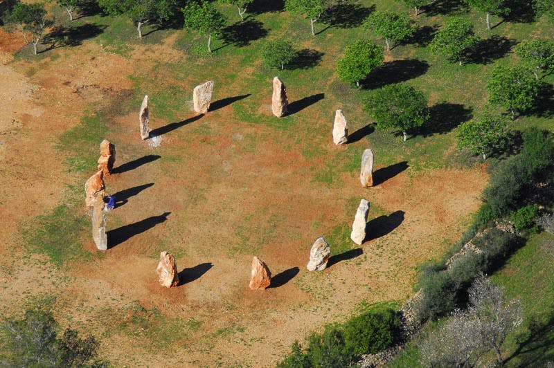 잔디와 나무로 둘러싸인 돌 원형 기념물의 공중 전망, 커다란 스탠딩 스톤이 그림자를 드리우고 있습니다.