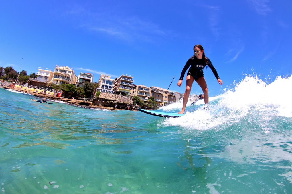 검은색 발진 보호대를 착용한 한 여성이 바위 해변의 건물을 배경으로 맑고 푸른 물결 속에서 서핑을 즐기고 있습니다.