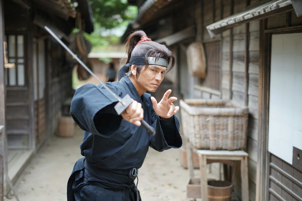 검은 전통 복장을 한 남성이 오래된 목조 마을의 경계 태세에서 칼을 들고 있습니다.