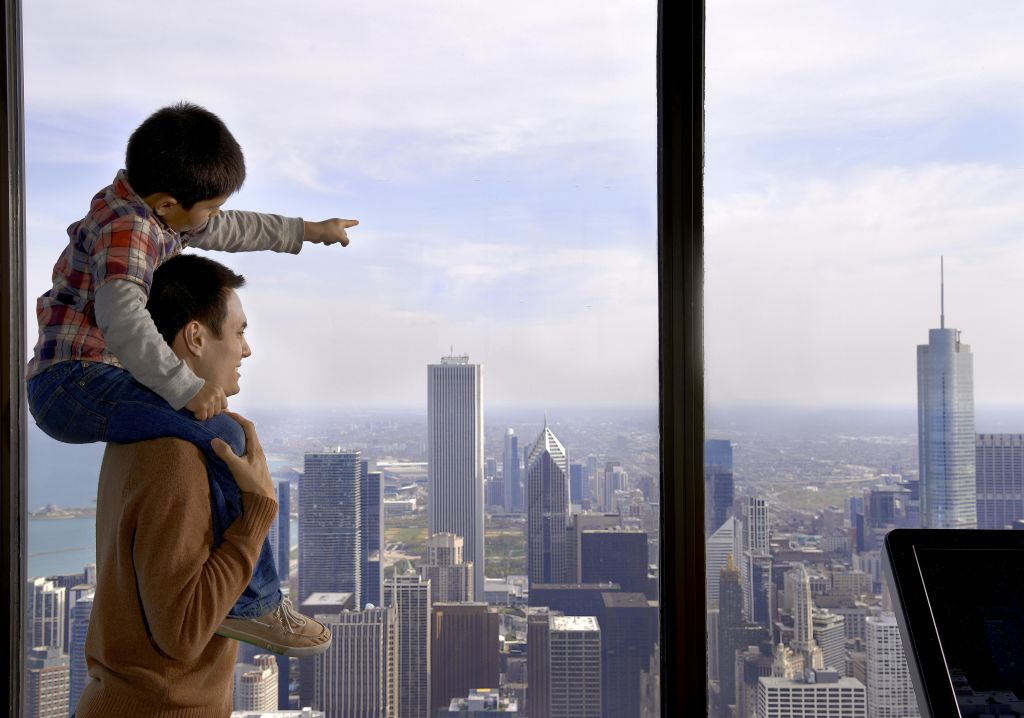 커다란 창문 너머로 보이는 도시의 스카이라인을 가리키는 한 남자가 아이를 어깨에 메고 있습니다.