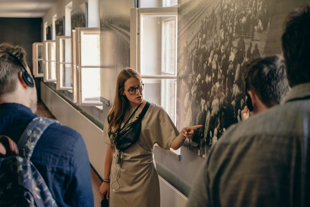창문과 벽에 걸린 역사적인 사진이 있는 박물관 복도의 한 그룹에게 설명하는 동안 가이드가 제스처를 취하고 있습니다.