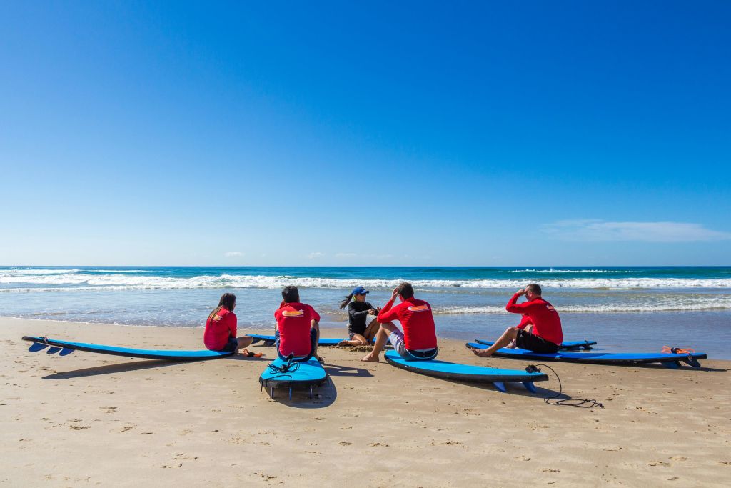 빨간 셔츠를 입은 5명이 서핑보드가 있는 해변에 앉아 맑고 푸른 하늘 아래 바다를 마주하고 있습니다.