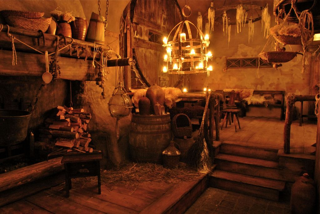 나무 가구, 바닥에 건초, 촛대 샹들리에, 쌓인 장작, 행잉 랜턴이 있는 소박한 방에 조명이 희미하게 켜져 있습니다.