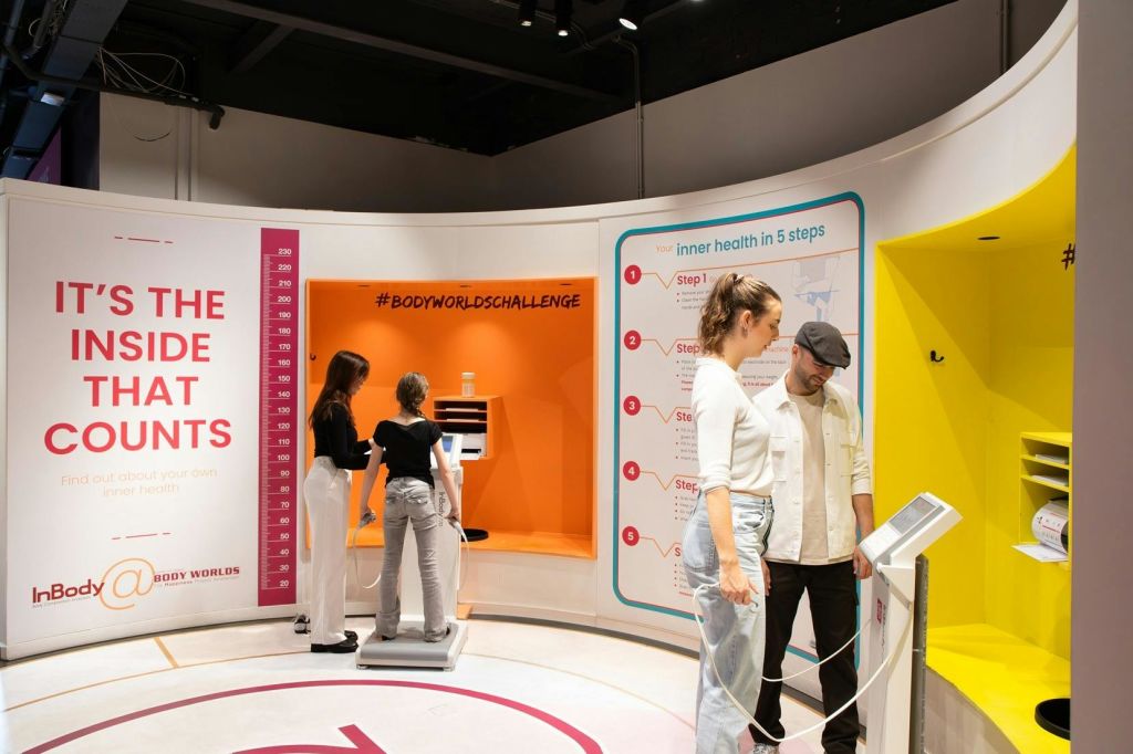 건강 단계 지침이 포함된 # BodyWorldsChallenge라는 최신 디스플레이 영역에서 대화형 건강 전시회를 사용하는 사람들.