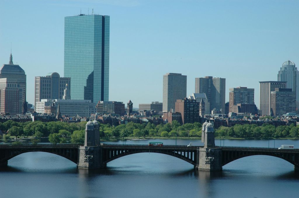 강을 건너는 다리, 높은 건물을 배경으로 푸른 하늘 아래 강둑을 따라 나무가 우거진 도시 풍경.