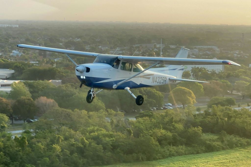 해가 질 무렵 숲 위를 날고 있는 작은 파란색과 흰색의 프로펠러 비행기로, 비행기의 등록 번호 N4325R이 보입니다.