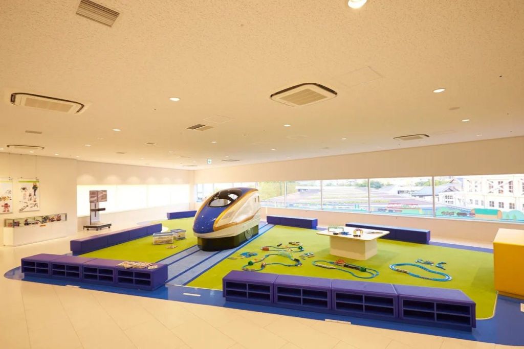 기차 테마가 있는 어린이 놀이 공간으로, 모델 기차 엔진, 트랙, 녹색 바닥에 다채로운 좌석이 있습니다.