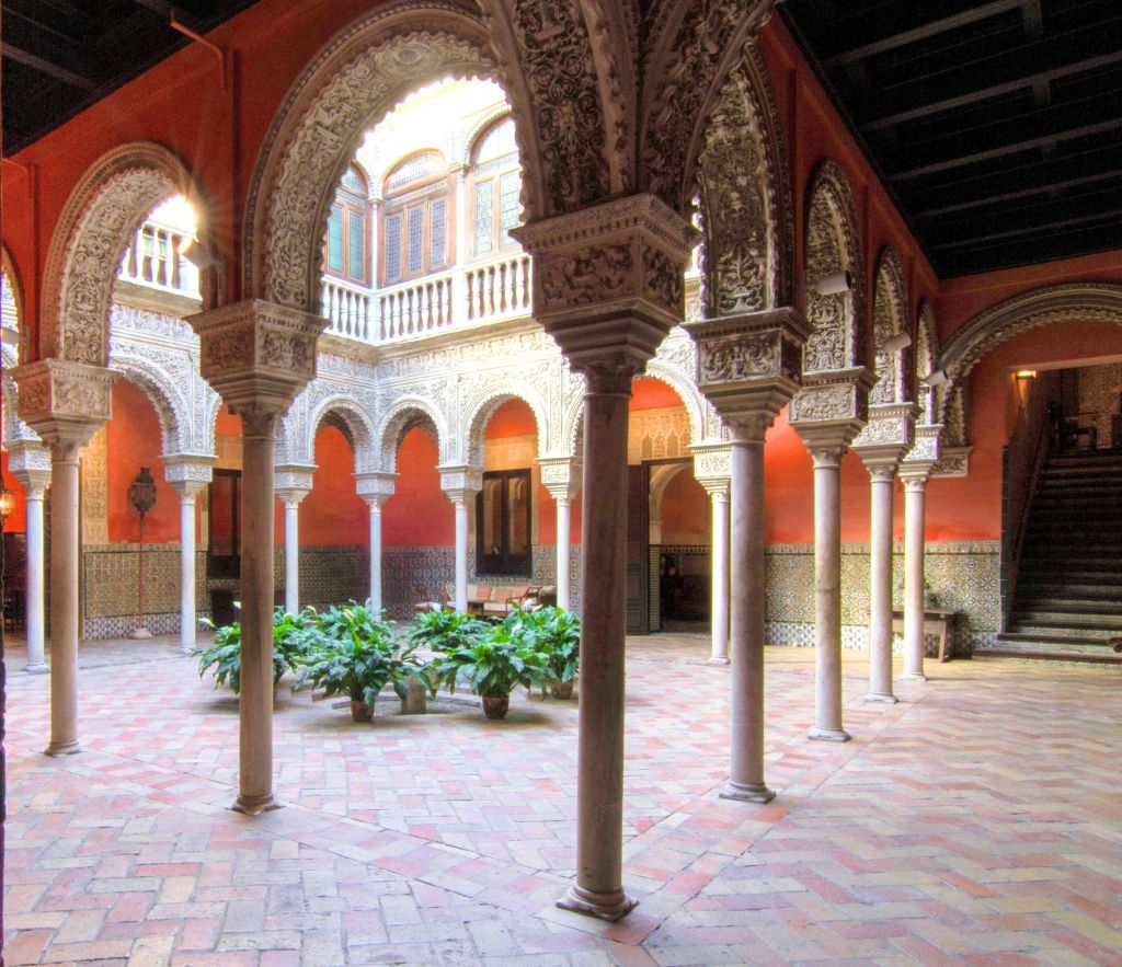 화려한 아치와 기둥, 타일 벽, 벽돌 바닥, 중앙에 화분이 있는 마당입니다.