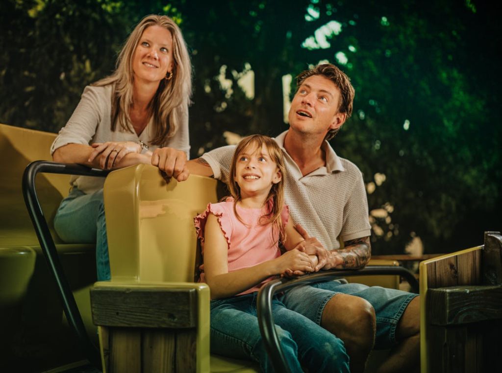 3인 가족이 단풍을 배경으로 놀이공원에 앉아 미소를 짓고 있습니다.